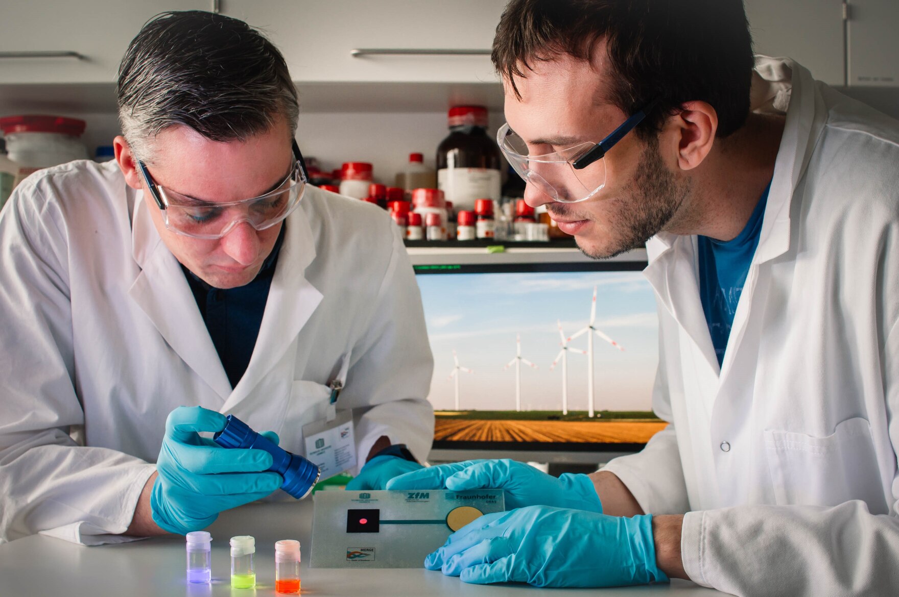Zwei Personen in weißer Schutzkleidung in einem Labor beleuchten ein Probestück mit einer Taschenlampe.